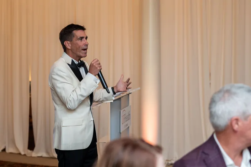 Person in cream jacket and bow tie holds a microphone at a podium, gesturing while speaking to seated guests in an elegant venue.