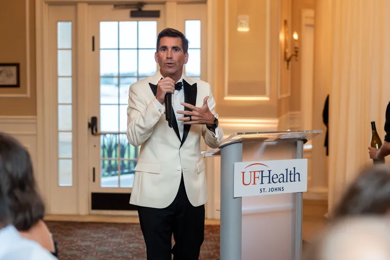 Speaker in cream blazer and black bow tie stands at a UFHealth podium with tall windows and river views behind.