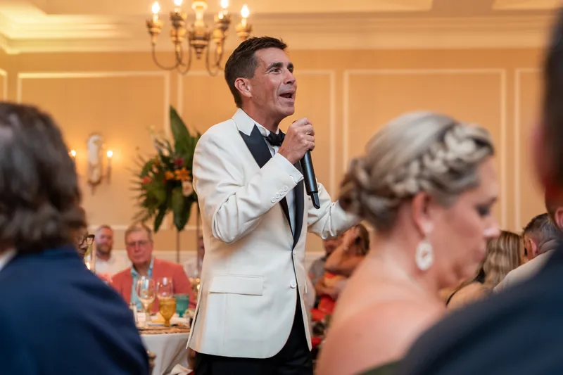Man in cream jacket and black bow tie addresses dining guests, smiling while holding a microphone at an elegant gala table.