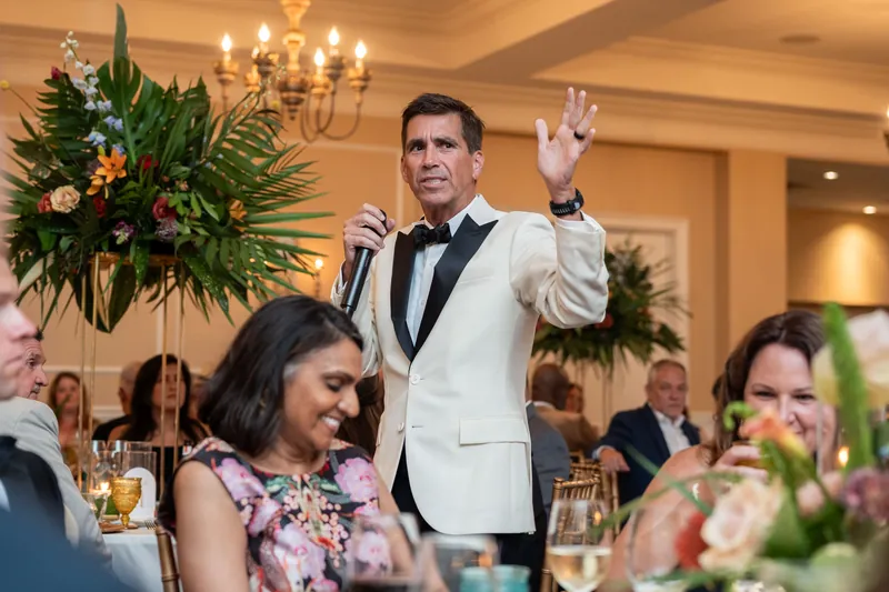 Person in cream tuxedo and black bow tie speaks animatedly among seated diners, flanked by tall floral arrangements.