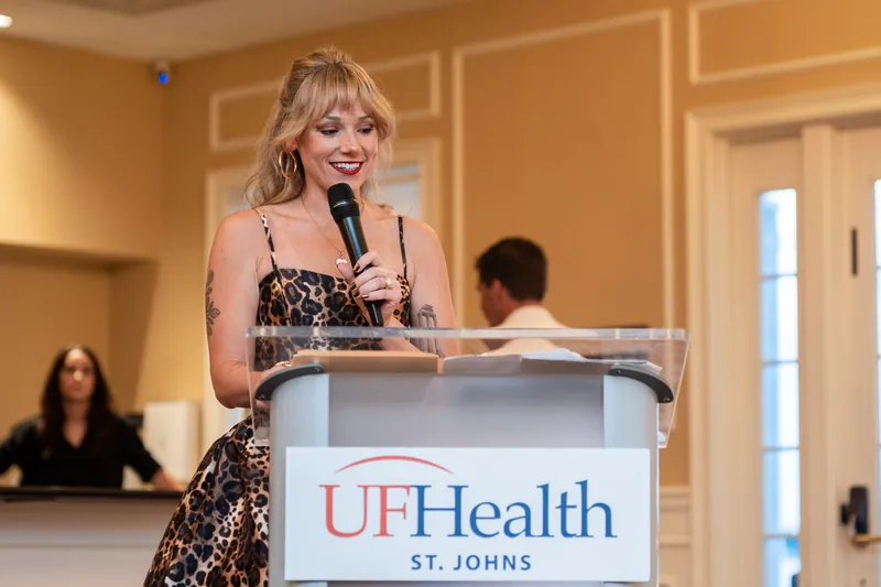 A woman in a sequined black gown speaks from a UFHealth podium in an elegant ballroom, with guests seated in the background.