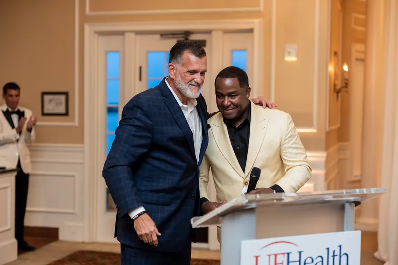 Two men in business attire smile together at a UFHealth podium inside a formal ballroom with white architectural details.