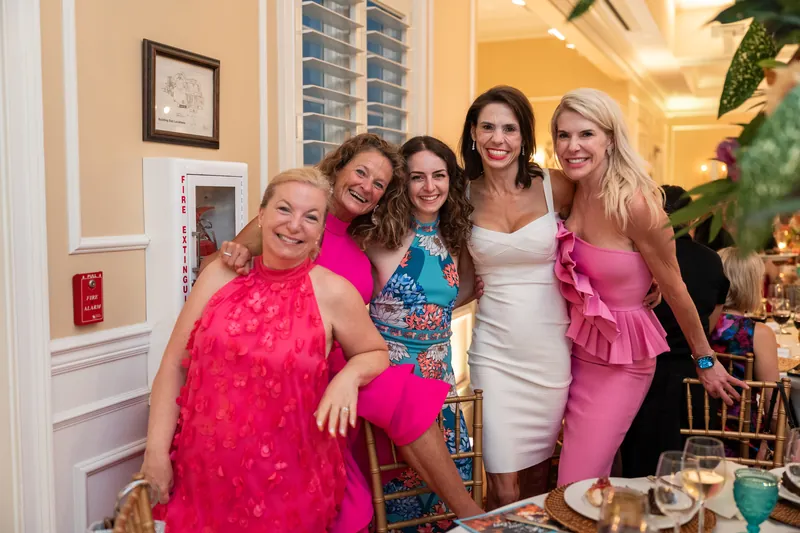 Five women in coordinated pink and jewel-toned gowns pose together indoors, smiling warmly at a gala event.