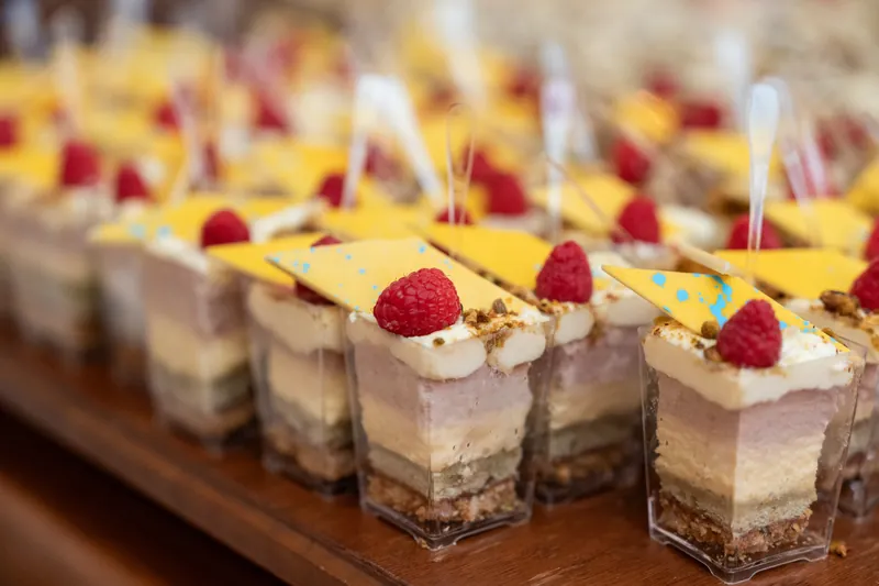 Tiered individual desserts with graham cracker bases, creamy filling, and fresh raspberries topped with yellow coulis line a wooden board.