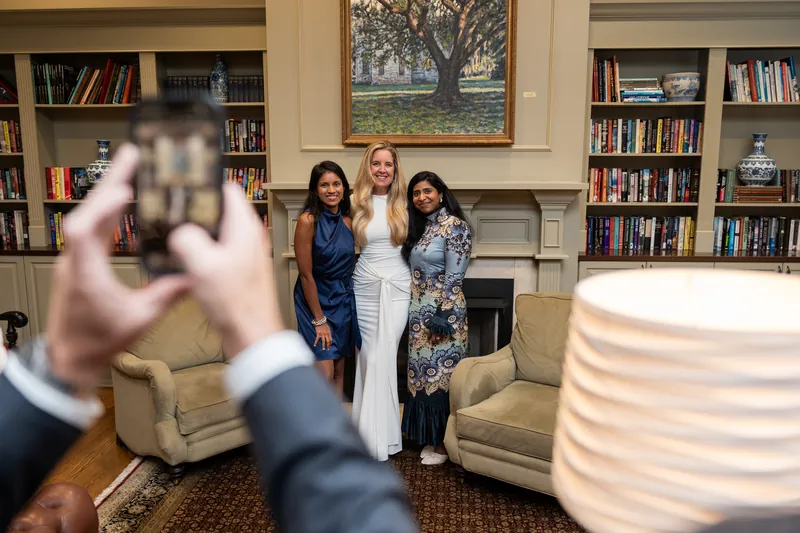 Someone in a suit holds up a phone to photograph three women standing together in River House Events' library, lined with bookshelves and warm lighting.