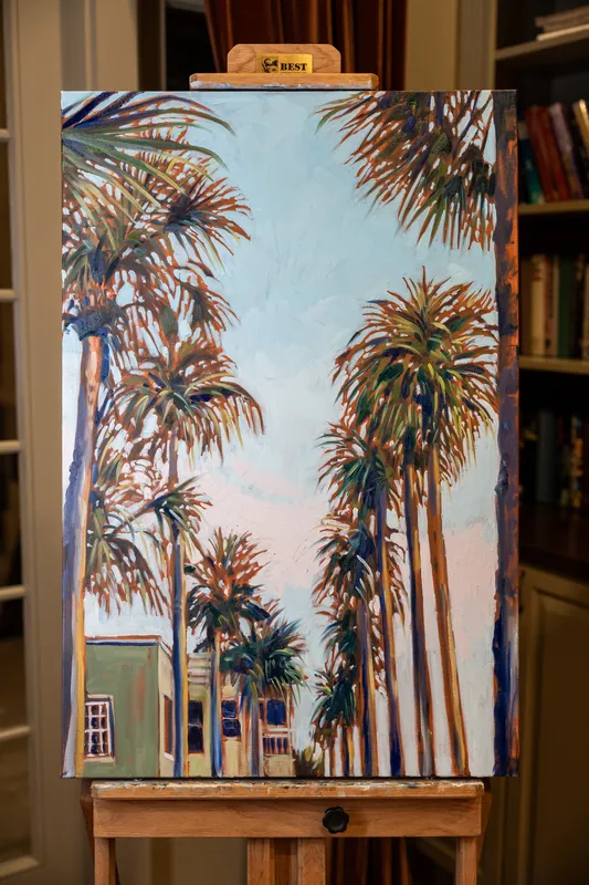 Framed painting of towering palm trees against a pastel sky sits on an easel between bookshelves in the venue's interior.