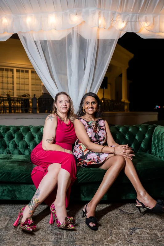 Two women in fuchsia and patterned dresses sit together on a green velvet sofa at River House Events.