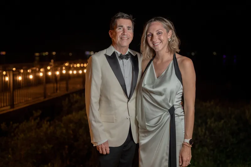 Two people in a beige blazer and sage gown stand on a lit deck overlooking the river at night.