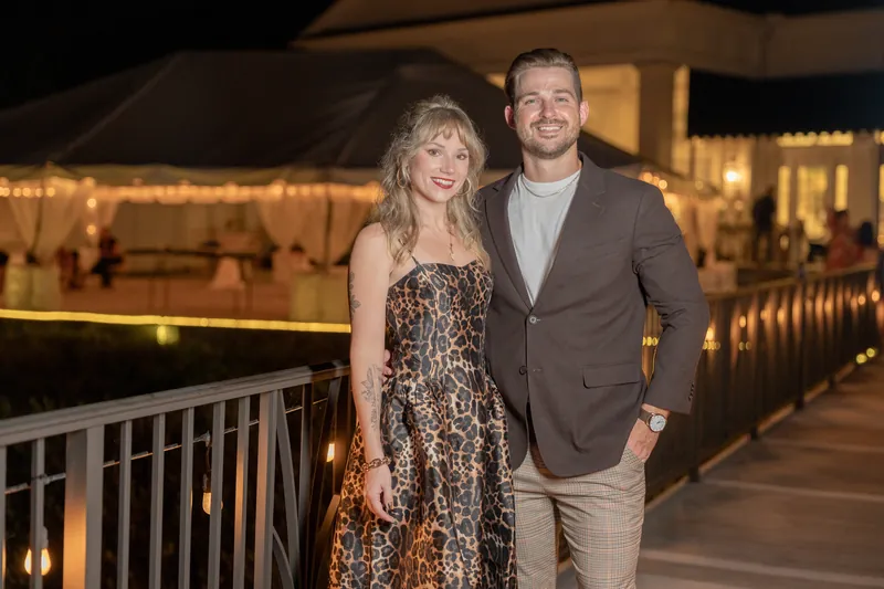 Two guests in a sequined dress and brown blazer stand together on a lit waterfront deck at River House Events, St. Augustine.