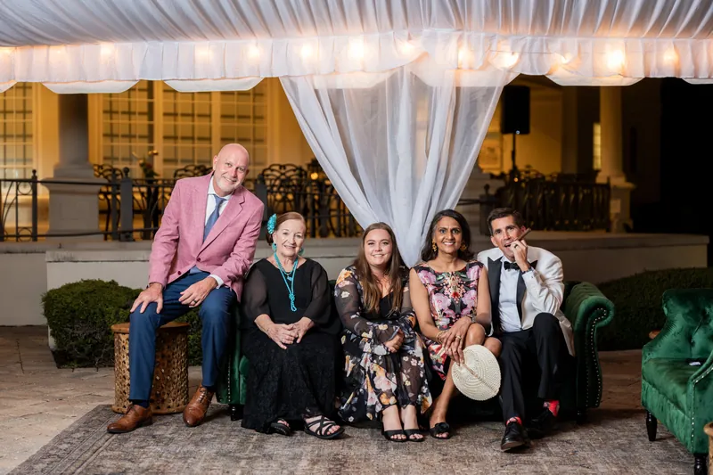 Five guests in formal wear sit on a green tufted bench under draped white fabric at the venue.