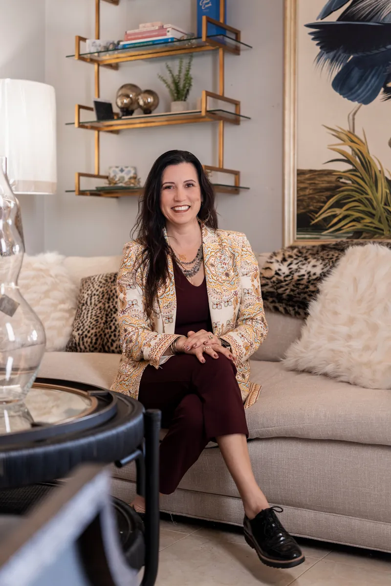 Natalie seated on a couch in a patterned blazer, smiling with styled showroom furniture and art behind her