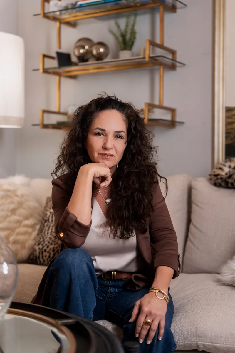 Valerie with a confident expression, chin on fist, seated on a couch with gold-accented shelving behind her