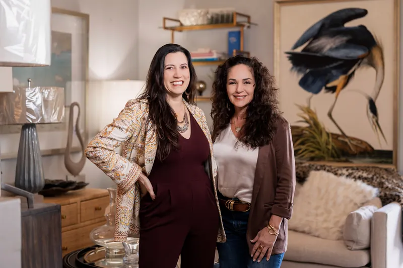Natalie and Valerie standing together with a great blue heron painting and brass shelving behind them