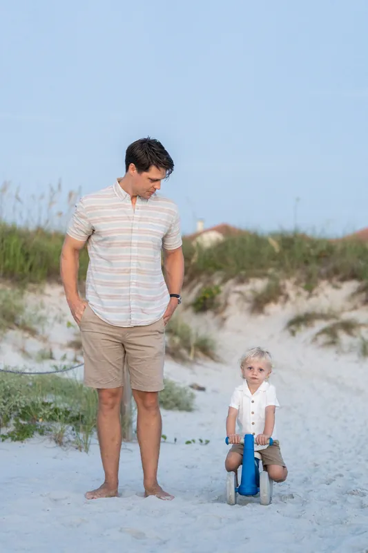 A man in a striped shirt and khaki shorts stands on the sand at St. Augustine Beach while a young child sits nearby