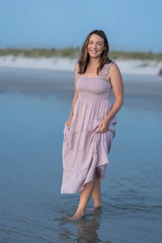 Jourdan walks through shallow water at St. Augustine Beach in a lavender dress, smiling as waves lap around her feet.