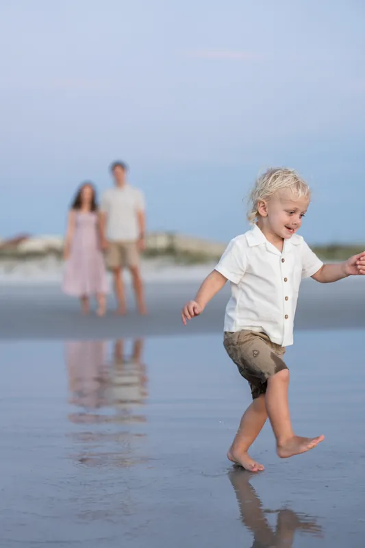 Blonde boy in white shirt kicks through shallow water while his family watches from behind at St. Augustine Beach.
