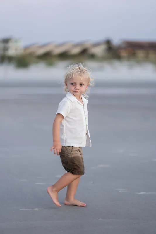 A blonde toddler in a white shirt and khaki shorts runs along the beach, looking back over his shoulder.