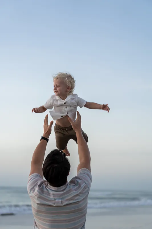 A man in a striped shirt lifts a small child overhead against the ocean backdrop at St. Augustine Beach.