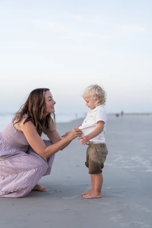 Jourdan crouches down to hold hands with a toddler in the shallow surf, both smiling at each other.