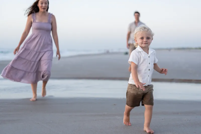 A toddler in a white shirt runs ahead on the sand while Jourdan follows behind in a lavender dress at St. Augustine Beach.