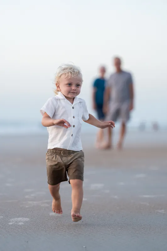 Blonde toddler in a white shirt and khaki shorts runs barefoot on the sand at St. Augustine Beach while family members stand
