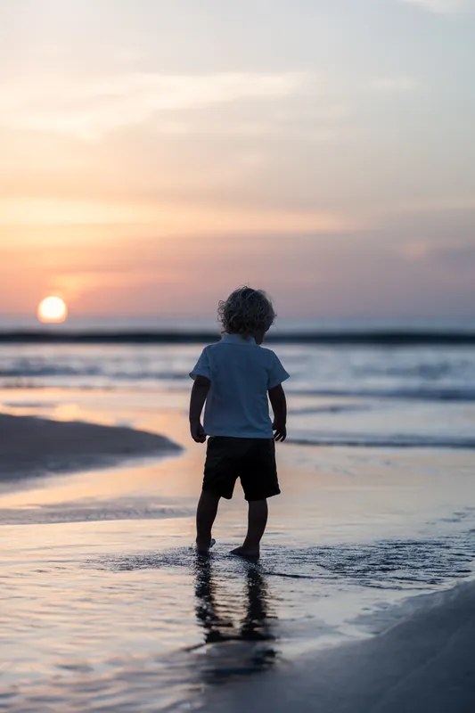 Child wearing a blue shirt stands in the shallow water at sunset, gazing toward the horizon as the sun dips low