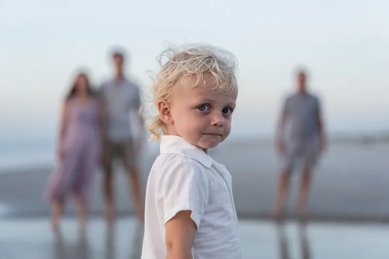 Young blonde boy in a white shirt looks over his shoulder at the camera while his family stands out of focus behind him