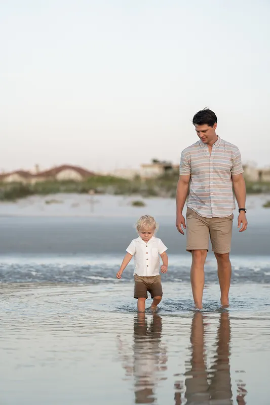 Man in a checkered shirt walks hand-in-hand with a toddler through the shallow surf at St. Augustine Beach.