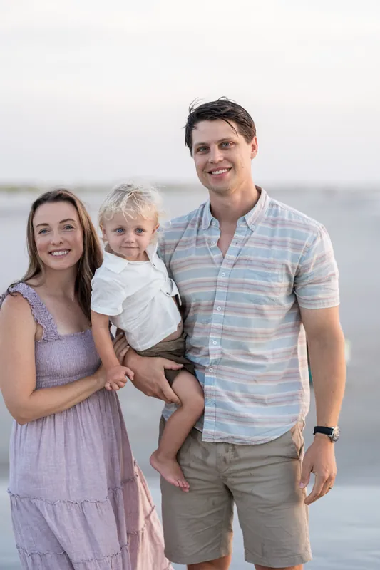 Jourdan and her husband smile at the camera while holding their young son, all three standing on the beach with the ocean