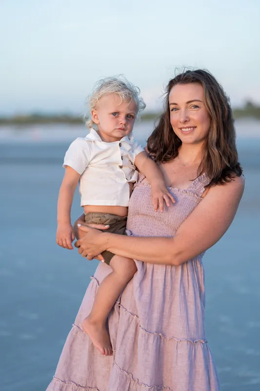 Jourdan holds her young son at St. Augustine Beach at dusk, both smiling toward the camera with calm water behind them.