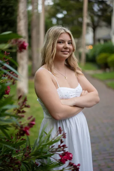 Zana stands with crossed arms in a white dress among flowering bushes on a brick pathway.