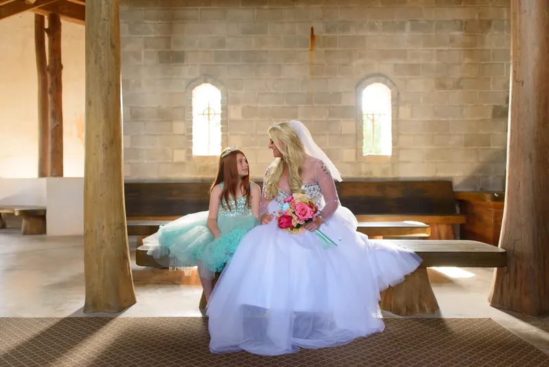 Bride and flower girl sitting on bench with matching tiaras and colorful bouquet in front of gothic windows at Castle Ottis