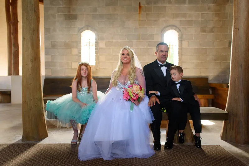 Family wedding portrait with bride, groom, flower girl, and ring bearer on bench in front of gothic windows at Castle Ottis