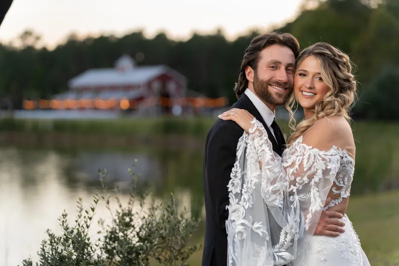 Couple embracing by lake with barn in background at sunset