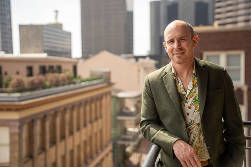 0008_Man in a green blazer standing on an urban balcony with tall buildings in the background.jpg