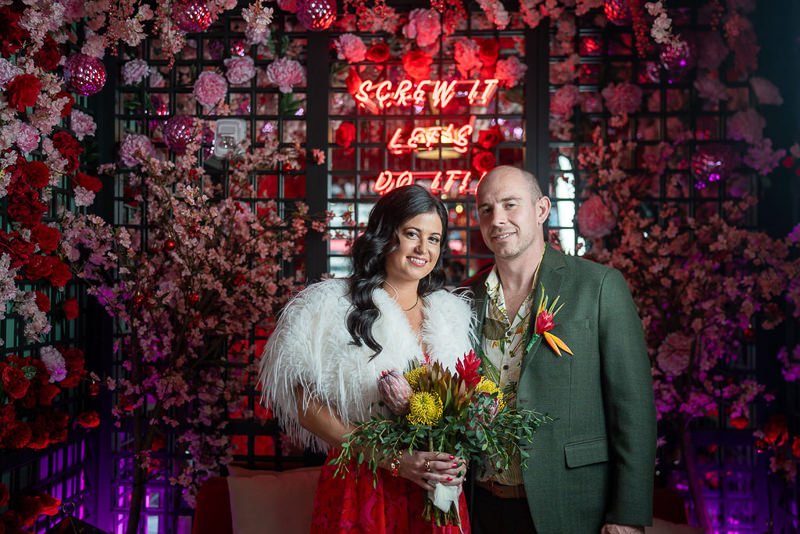 0014_A couple stands before a floral backdrop with a neon sign that reads Screw it let s do it.jpg