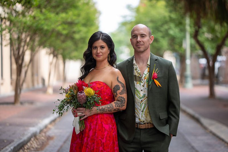 0015_A couple stands together on a tree lined street the woman in a red dress holding flowers and the man in a green suit with a tropical shirt.jpg