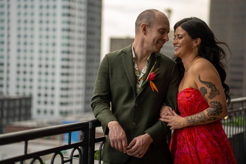 0019_A couple smiling and embracing on a city rooftop with the man in a green suit and the woman in a red dress.jpg