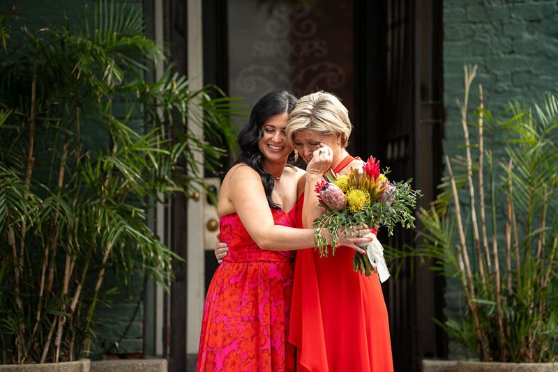 0022_Two women in red dresses share an emotional moment with one holding a bouquet of flowers.jpg