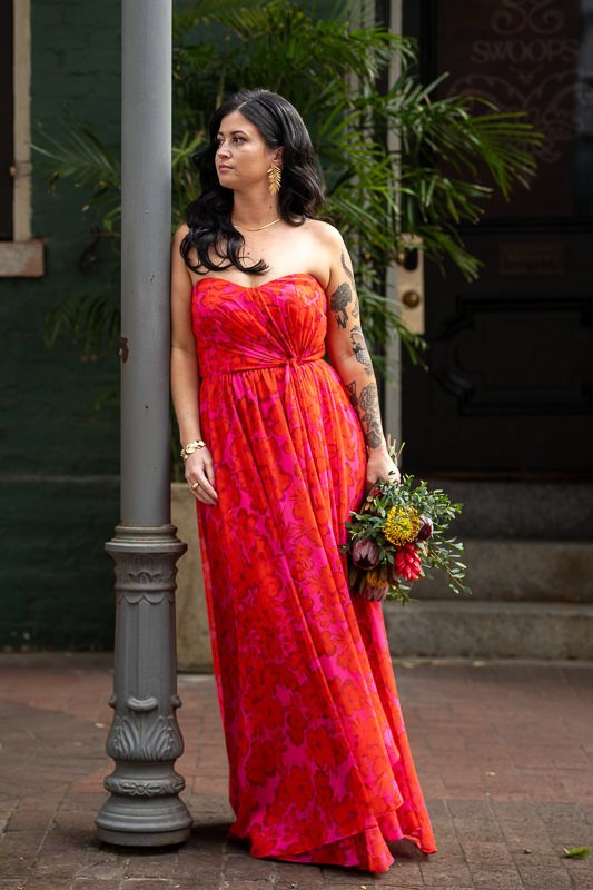 0023_Woman in a vibrant red orange strapless dress holding a bouquet stands leaning against a lamppost gazing to the side.jpg