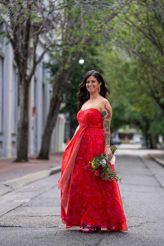 0024_Woman in a vibrant red dress holds a bouquet and smiles while standing on a tree lined street.jpg