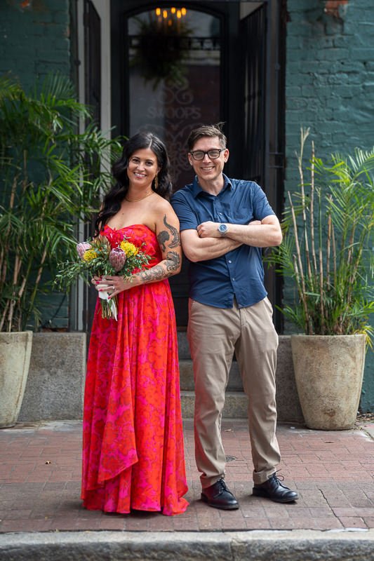 0025_A woman in a bright red dress holding flowers stands next to a casually dressed man in front of an entrance.jpg
