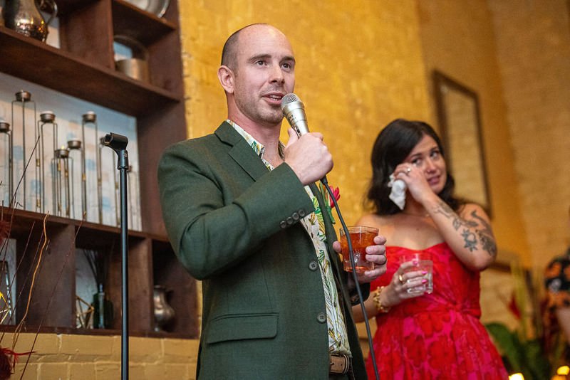 0028_A man addresses the crowd with a microphone while a woman in a red dress stands beside him holding a tissue and a drink.jpg