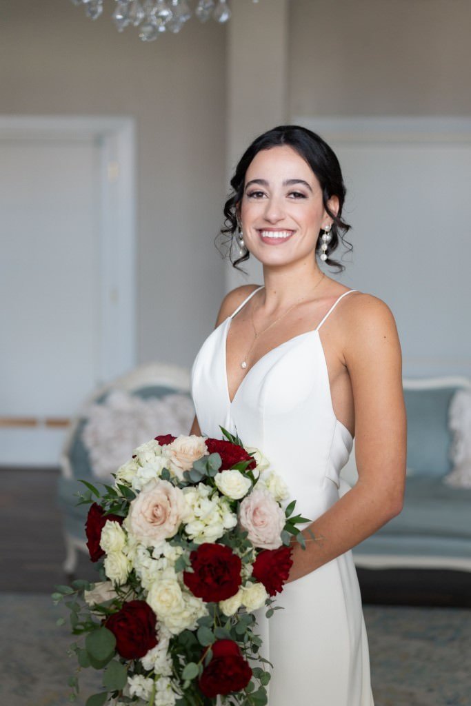 A bride in a white dress smiles while holding a bouquet of red and white flowers.jpg