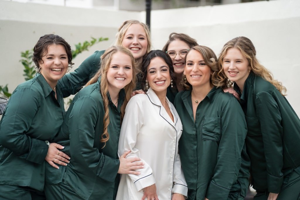 A bride in white pajamas and bridesmaids in green pajamas smile together.jpg