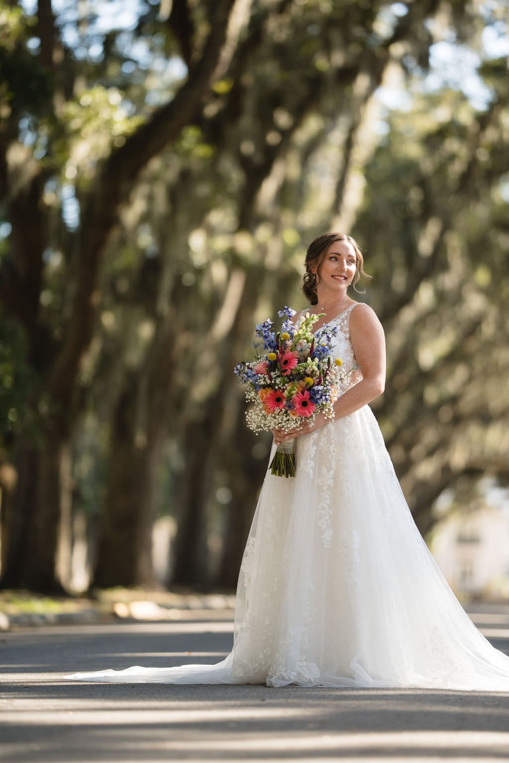 A bride stands in a beautiful tree lined street holding her bouquet and looking towards the sun She s ready for her big day.jpg