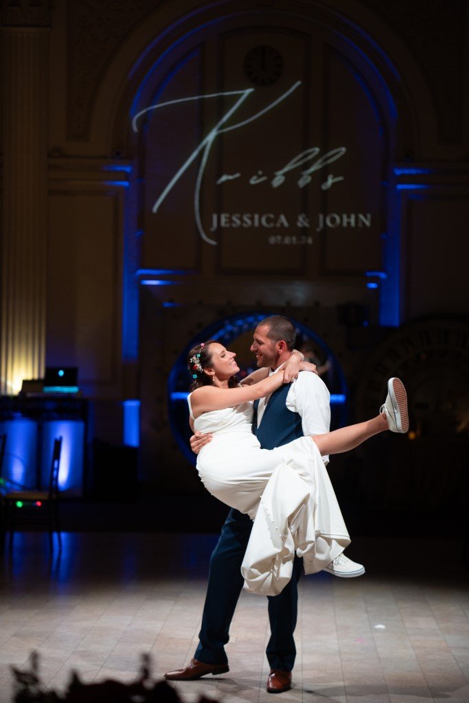 A couple shares a joyful dance under a spotlight with their names Jessica John illuminated in the background.jpg