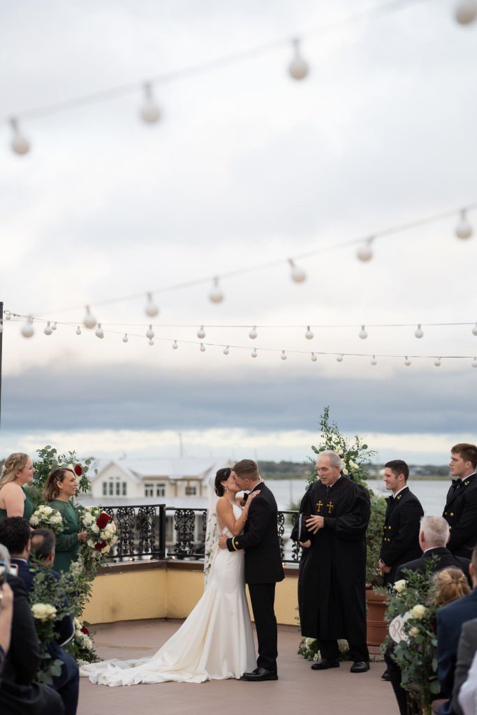 A couple shares their first kiss as newlyweds surrounded by friends and family under a string of lights.jpg