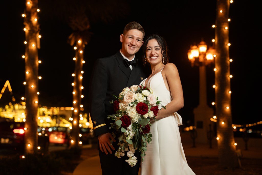 A joyful couple poses together under twinkling lights holding a beautiful bouquet during an elegant evening celebration.jpg