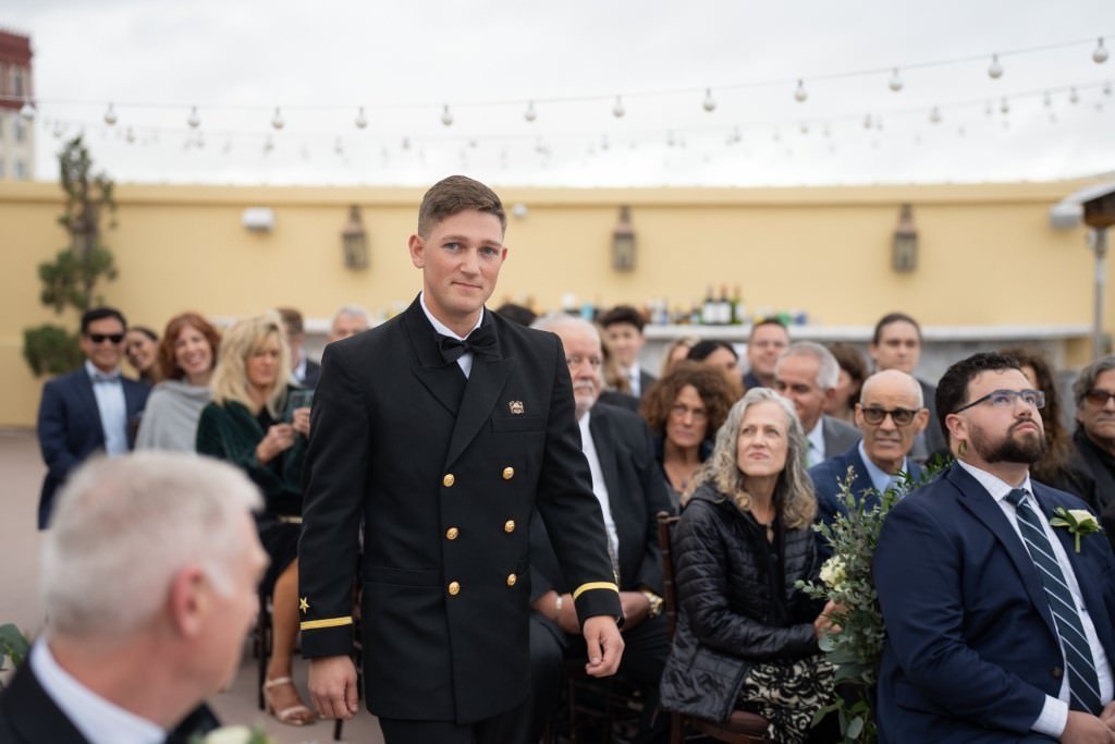 A man in a naval uniform walks down an aisle at an outdoor event surrounded by seated attendees.jpg